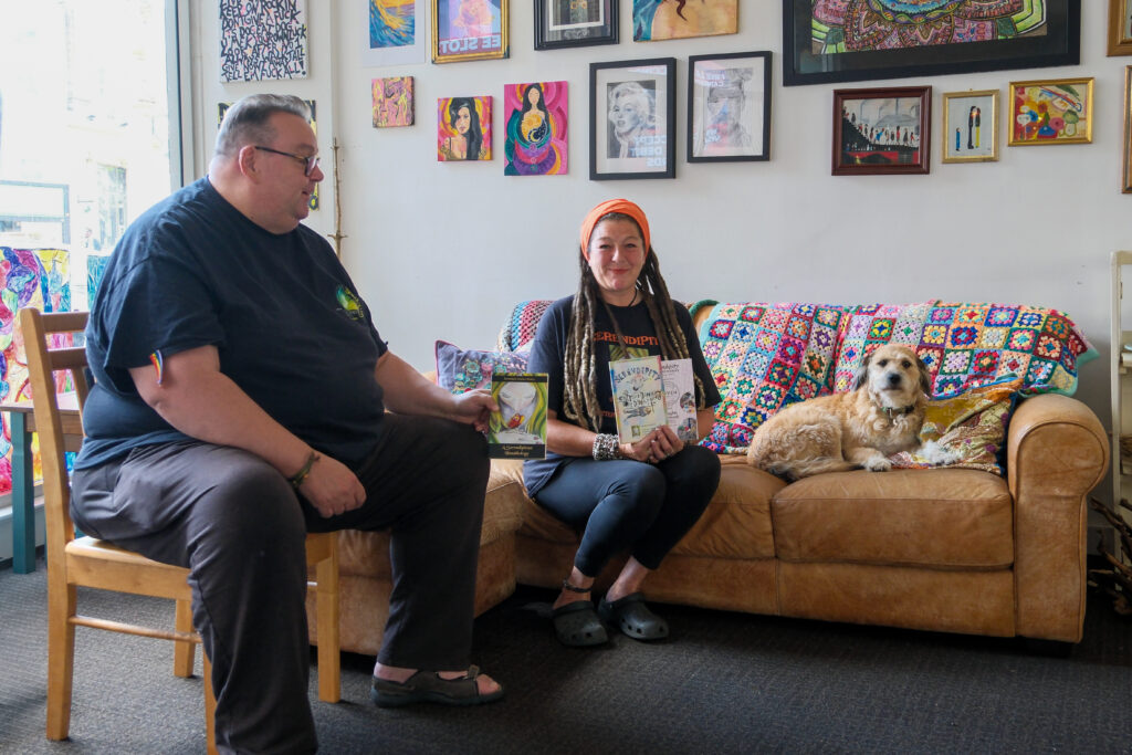Sonia and Martin holding book anthologies and sat on sofa with a dog.
